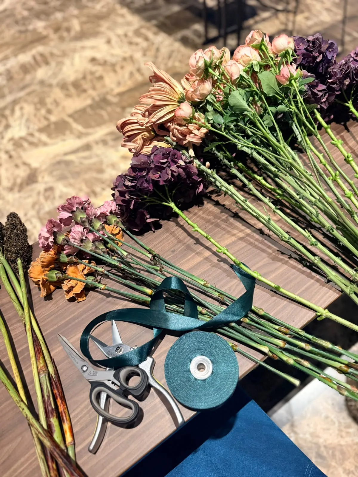 Fiori sul tavolo di lavoro: ortensie prugna, rose cipria, garofani ramati, forbici e nastro verde petrolio